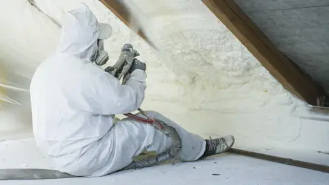 Getty Images Craftsman sits on the floor of an attic and uses tools to apply spray foam insulation to the underside of the roof. He is wearing white overalls and a respirator mask