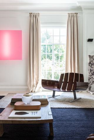 Blue and white living room with a neon pink painting next to the window on the back wall. There is an oversized wooden chair and a wooden coffee table in front of the window.