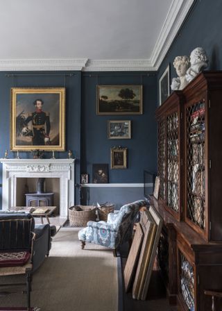 A living room painted dark blue. There are several traditional paintings hanging on the wall, a white marble fireplace and a large wooden chest full of books on the right side of the picture