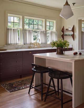 A bright and natural kitchen with plum purple kitchen cabinets and a kitchen island painted in a dusty pink shade. The countertops are made of white marble and the windows have half curtains