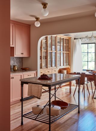 Shot of an open kitchen leading into a cozy dining room. The cupboards are painted dusty pink and there is a small, open kitchen island in the middle of the room.