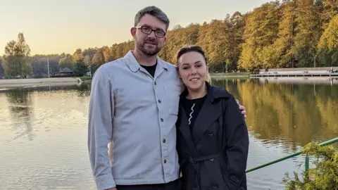 Robbie Anderson Robbie Anderson stands in front of a lake with his wife. He is wearing a gray button-up corduroy jacket and glasses and has his arm around his wife, who is wearing a dark gray trench coat with a belt.