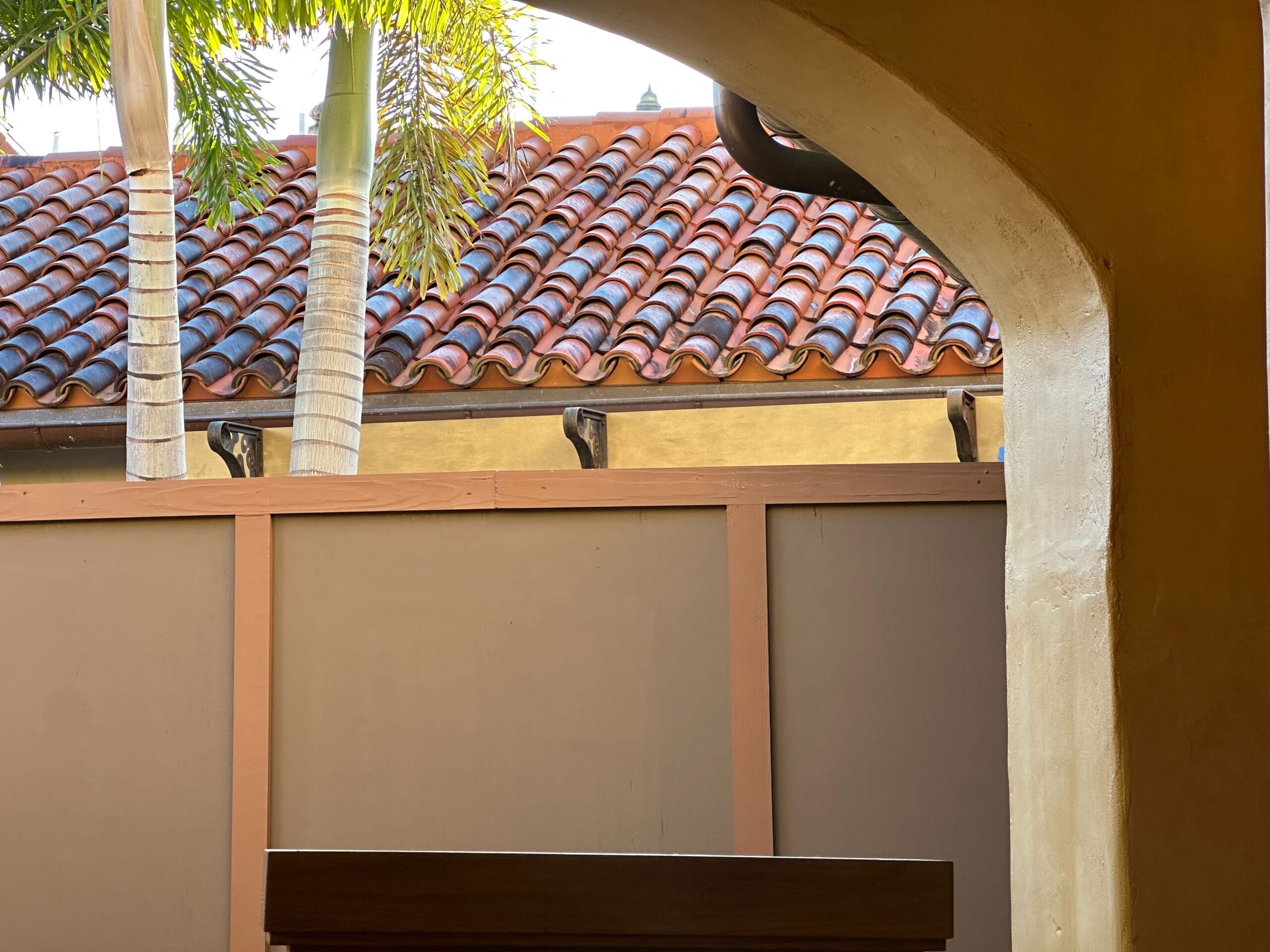 View of a Spanish-style tiled roof behind a wooden fence framed by an archway and palm trees.