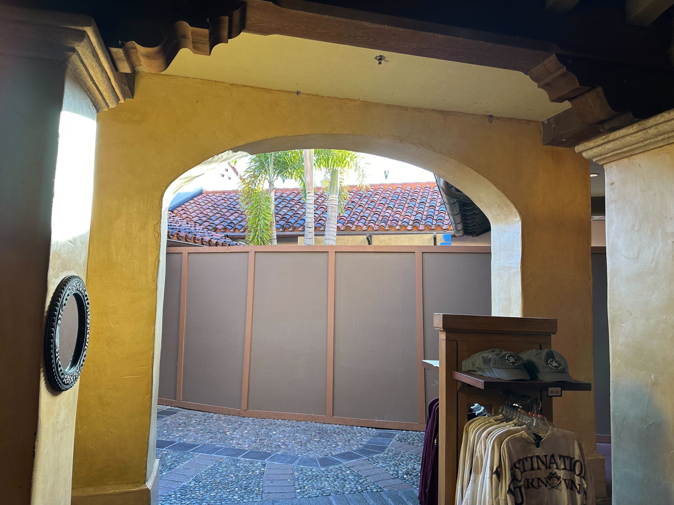 A view through an arched doorway showing a building wall outside. Inside you can see a shelf with shirts and hats.