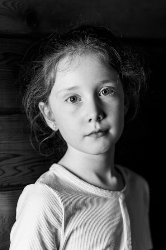 Black and white portrait of a young girl with light hair wearing a light top. She stands against a wooden wall and looks directly into the camera with a neutral facial expression. The lighting emphasizes her facial features.