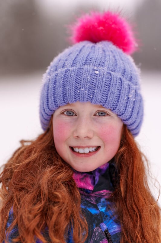 Smiling young girl with red hair, wearing a purple knitted hat with a pink pom-pom and a colorful winter jacket, standing in a snowy environment. Snowflakes decorate her hat and her cheeks are rosy.