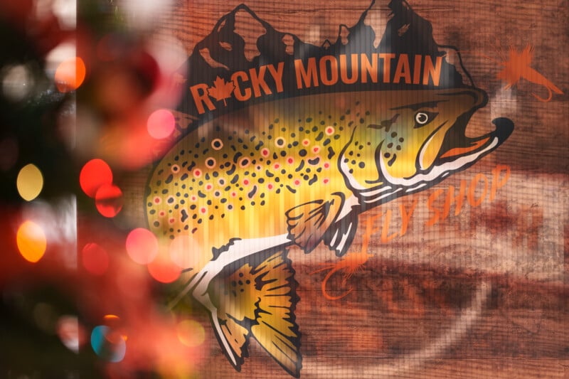 A festive scene shows a blurred Christmas tree with colorful lights on the left. On the right there is a painted sign with a trout fish and text that reads "Rocky Mountain Fly Shop" against a wooden background.