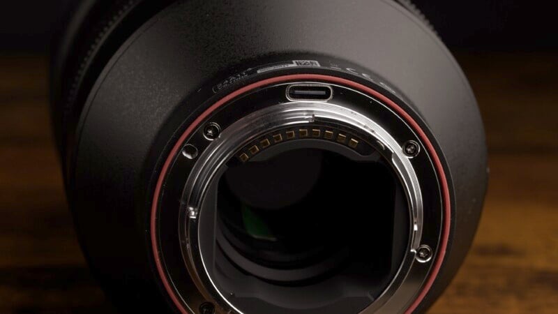 Close-up of the rear mount of a camera lens, highlighting the metal contacts and circular opening. The outer ring has a red band and the background is dark, highlighting the lens details.