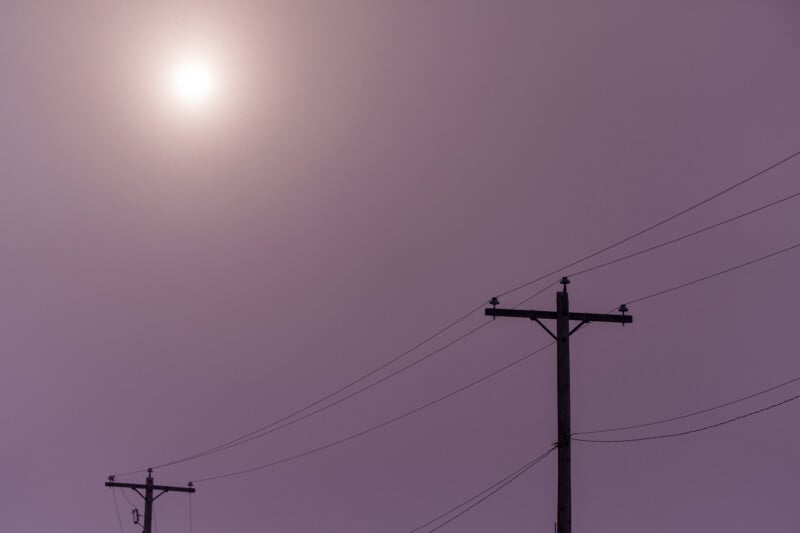 Two silhouettes of power lines and poles against a hazy, purple colored sky with a faint sun in the background, creating an ethereal and minimalist scene.