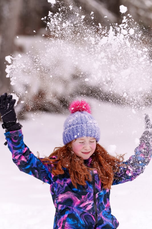 A child in a colorful winter jacket and blue knitted hat with a pink pom-pom happily throws snow into the air. The background is a snowy landscape with blurred trees. The child has long red hair and is wearing black gloves.