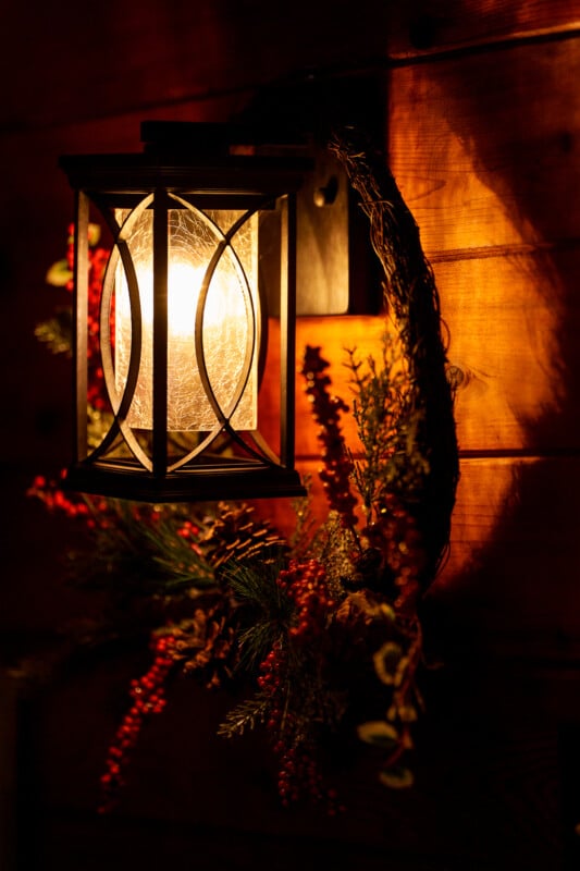 A softly glowing wall lantern with a decorative glass cover, mounted on a wooden wall. A festive wreath is decorated with red berries and green plants around the lantern, spreading warm light and creating a cozy atmosphere.