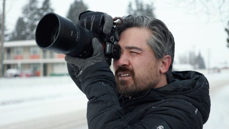 A person in a black jacket and gloves holds a camera in front of his face and takes a photo. You are outside on a snowy day with trees and a building in the background.