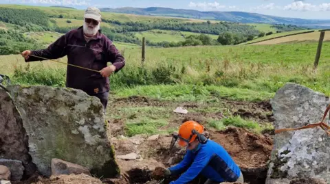 LDNPA Two people work to measure the slate shards and place them in the ground, which is then dug up. The site is located in an agricultural area in Jackson Ground, Lake District.