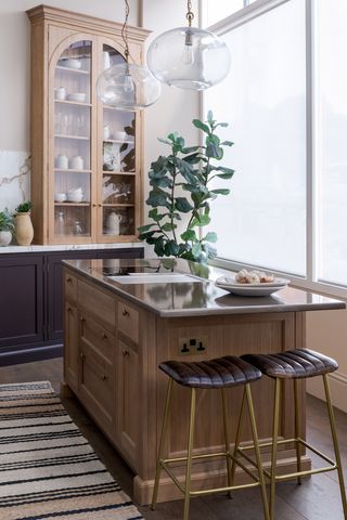 Beige kitchen with a light wood kitchen island. In the corner are two gold and brown leather bar stools, a striped rug and a large green plant. The island has a lava stone countertop and there are two clear pendant lights above the counter