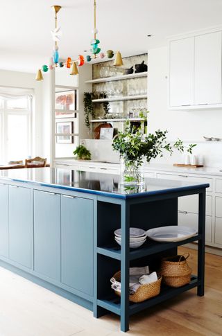 An all-white kitchen with hardwood floors and a dark blue kitchen island. The island has a lava stone countertop and a clear vase with green stems sits on the countertop. A colorful light hangs over the island.