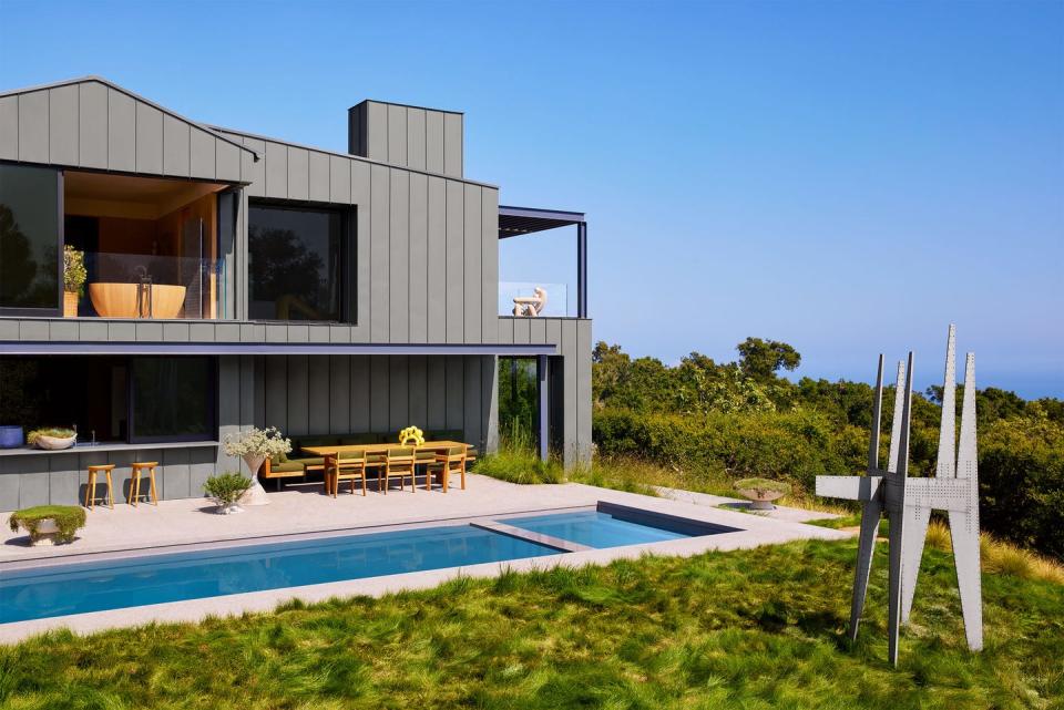 On the 2nd floor at the back of the house there are two windows, one of which shows a bathtub, and a terrace. Outside the 1st floor is a dining table and chairs next to a built-in lap pool and a metal sculpture