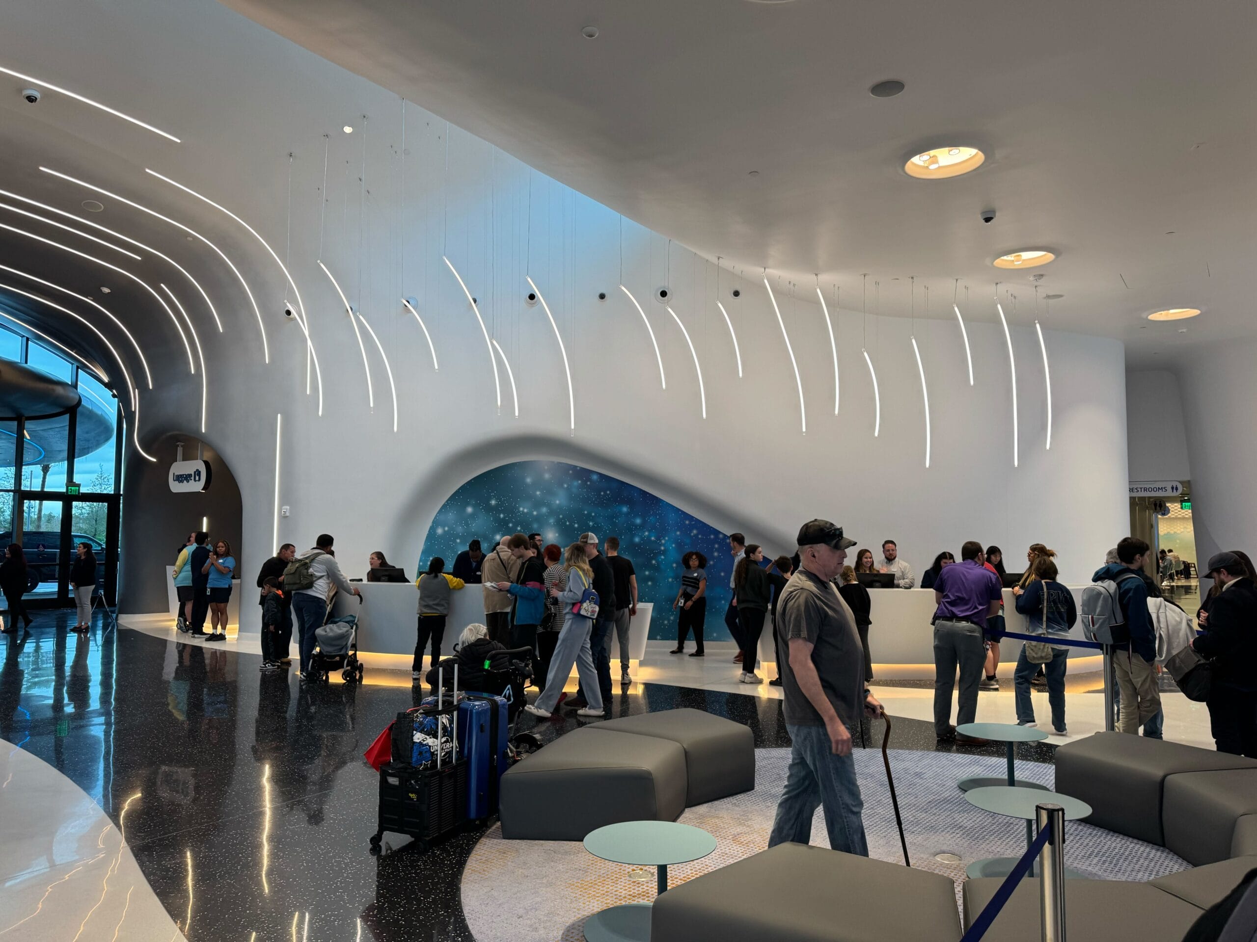 People queue at a modern hotel reception with a curved design, decorative lights and a blue accent wall. Luggage is visible near the seating area.