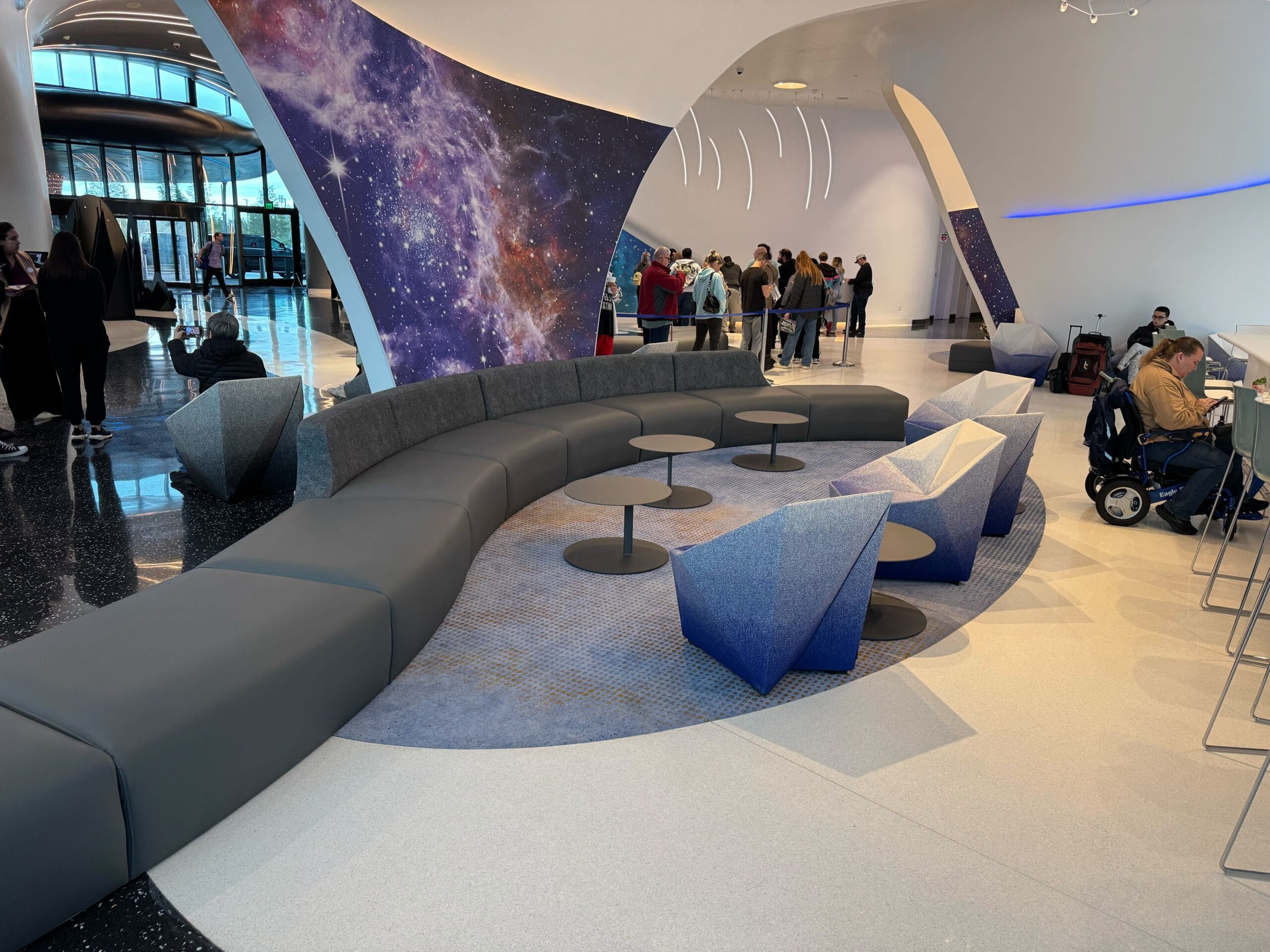 Modern lobby with a curved gray sofa, abstract geometric chairs and galaxy-themed wall art. In the background, people are gathered at a counter.