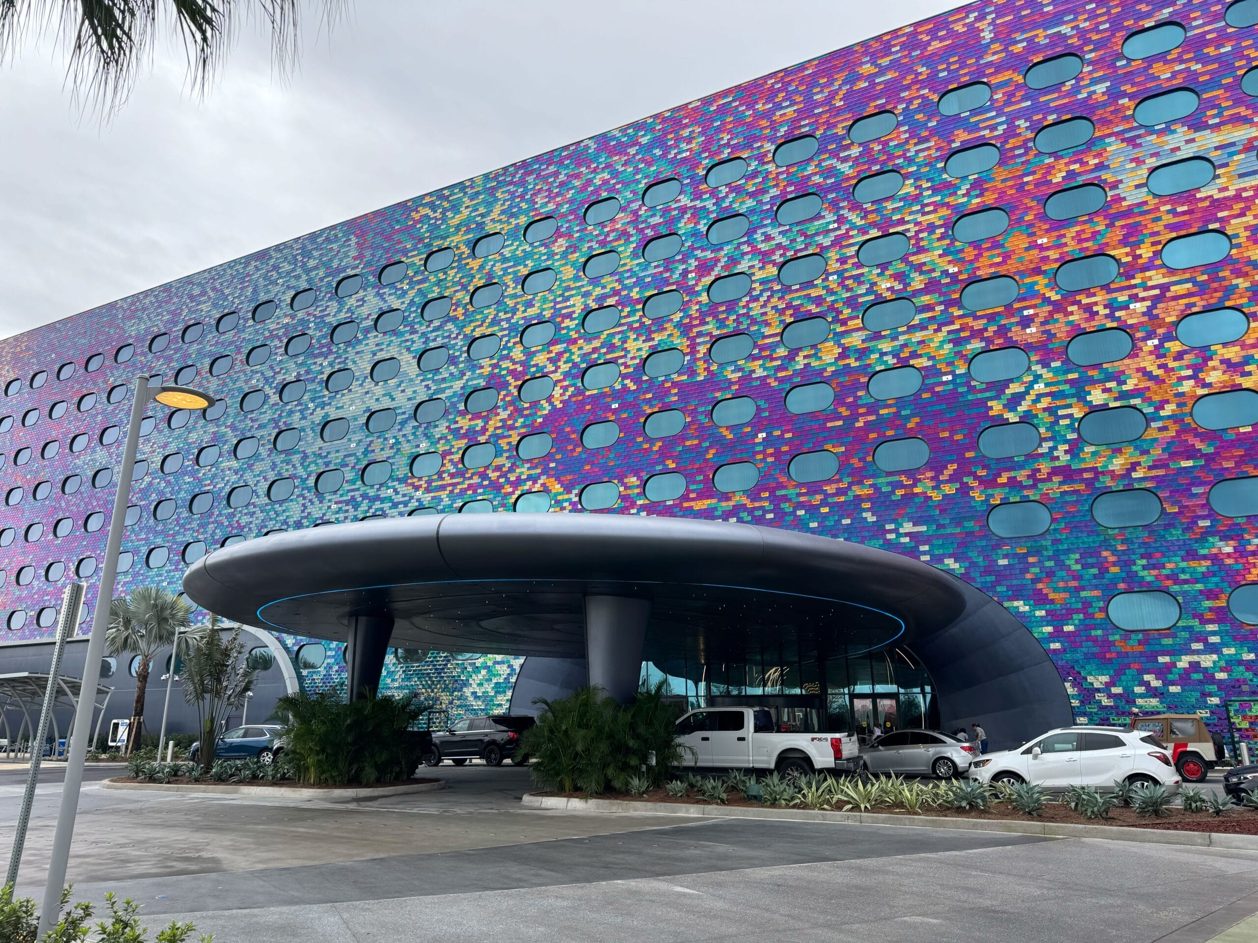 Colorful modern building facade with circular patterns over a round entrance. There are several vehicles and palm trees near the entrance. The sky is overcast.