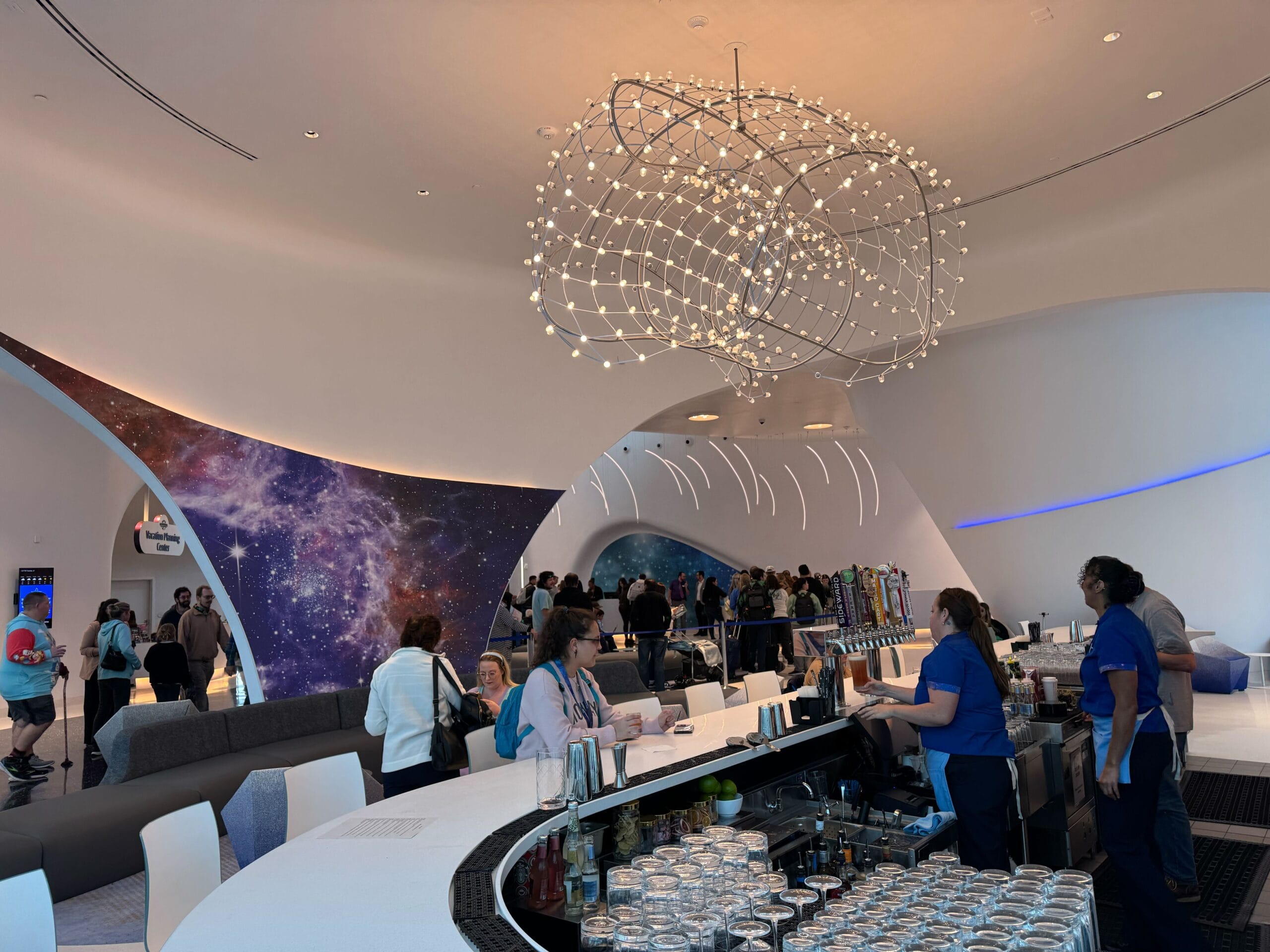 People gather in a modern bar with futuristic decor, a large abstract light fixture, and a cosmic-themed mural.