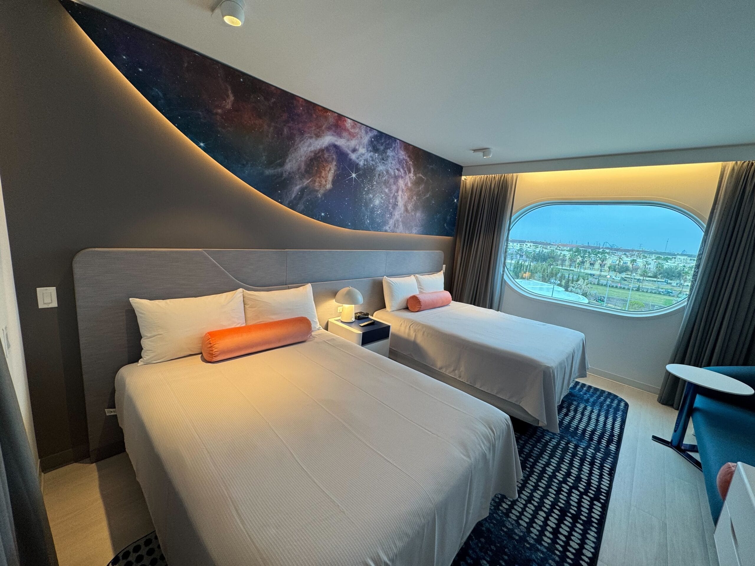 Hotel room with two neatly made beds, orange pillows and a space-themed mural. The large oval window with curtains offers a picturesque view.