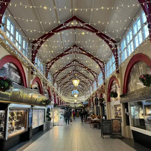 The inside of the market. Fairy lights hang above the hall lined with shops.