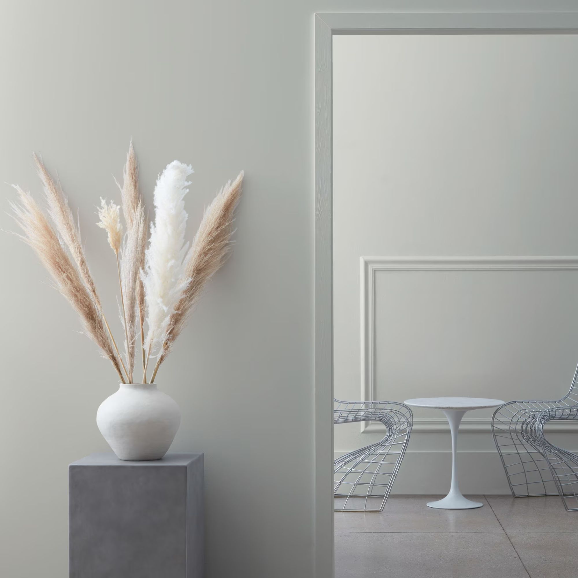 Light gray wall with base with pampas leaves, doors in gray room with metal chairs and round white table