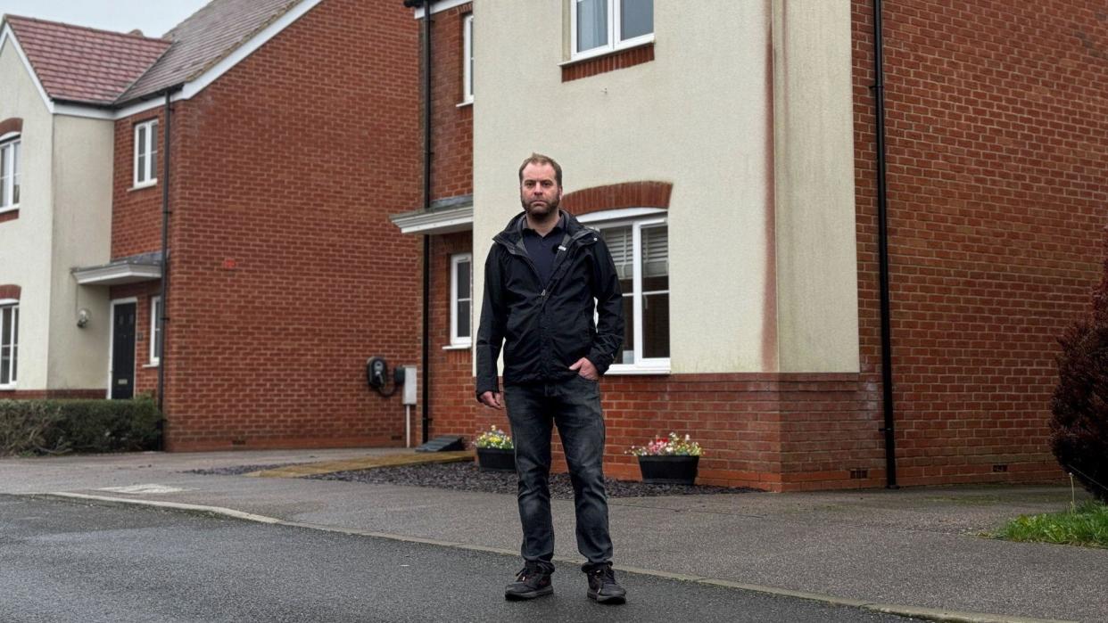 Dean Carpenter stands in front of his newly built house and has a large dark red stain on one corner of the walls