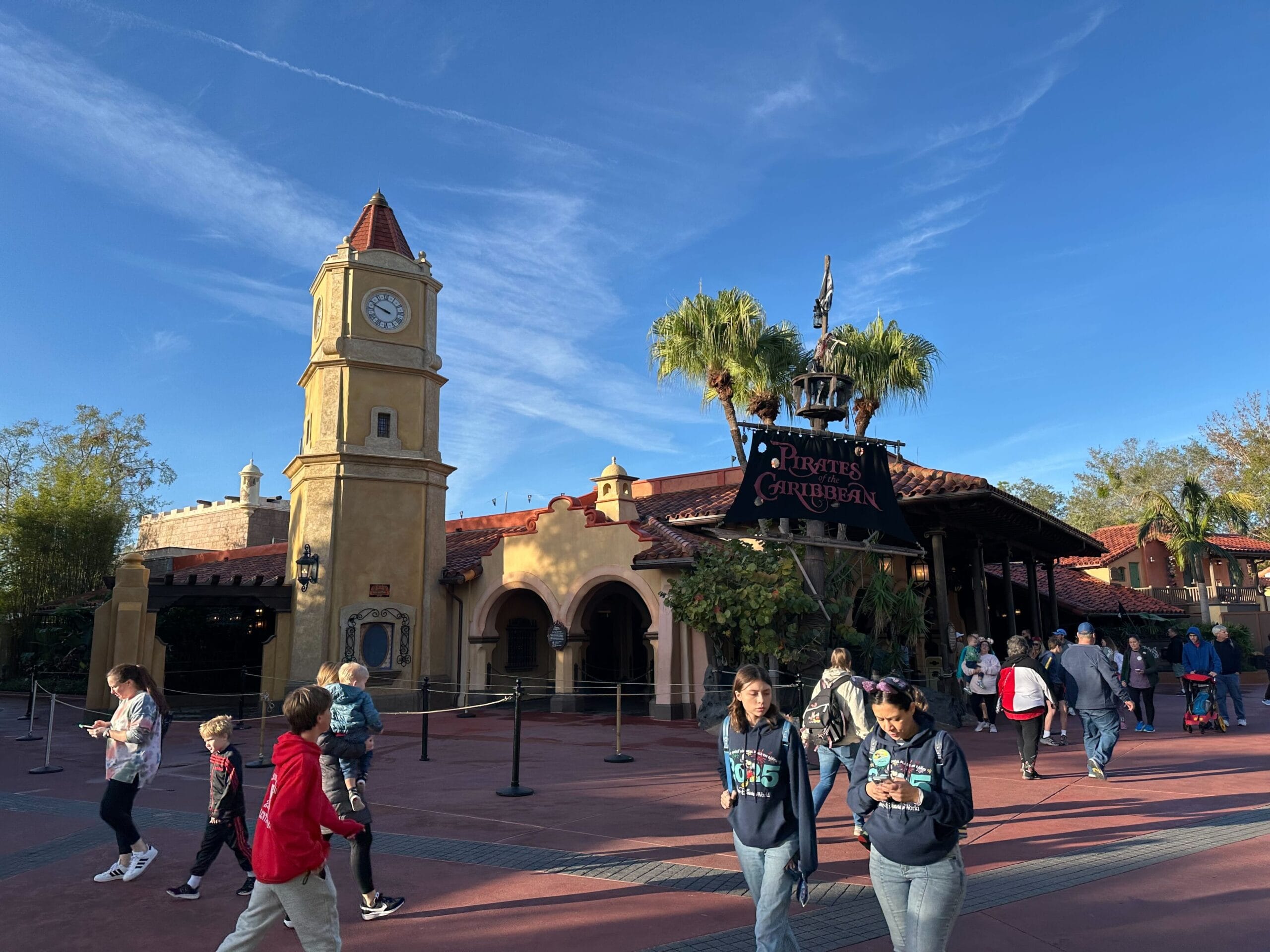 People walk near the entrance of the "pirates of the Caribbean" Attraction with a clock tower and tropical plants under a clear blue sky.