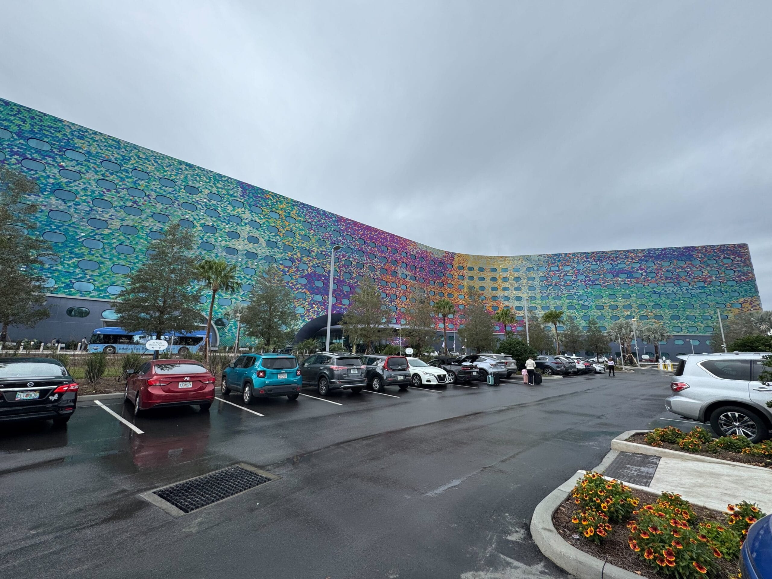 Colorful building facade with a honeycomb pattern, adjacent to a parking lot with cars. Overcast sky and surrounding greenery visible.