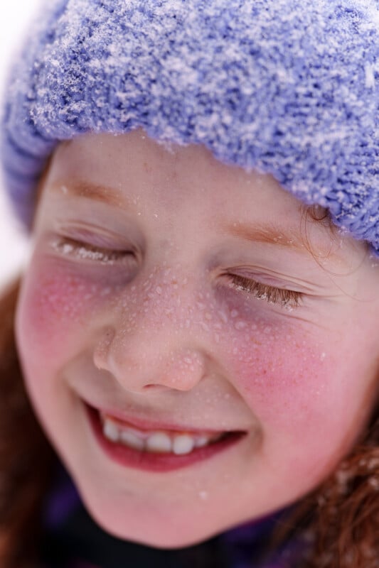 A close-up of a smiling child with eyes closed and wearing a blue knitted hat covered in snowflakes. The child's cheeks are rosy and there are tiny drops of water on the eyelashes and skin.