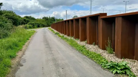 Charles Clover a street that flows through the landscape with a high brown steel wall on the right side