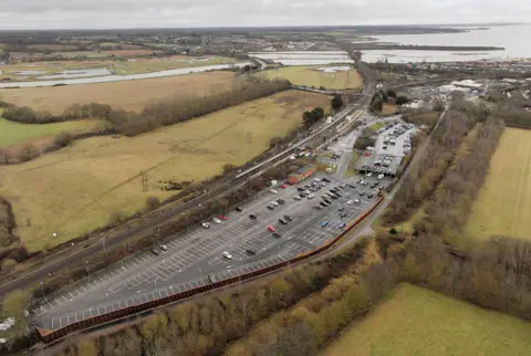 Stephen Huntley/BBC Aerial View of the Manningree train station, which shows a large parking lot along the railway line. A river and a mouth can be seen behind it, together with fields and other buildings.