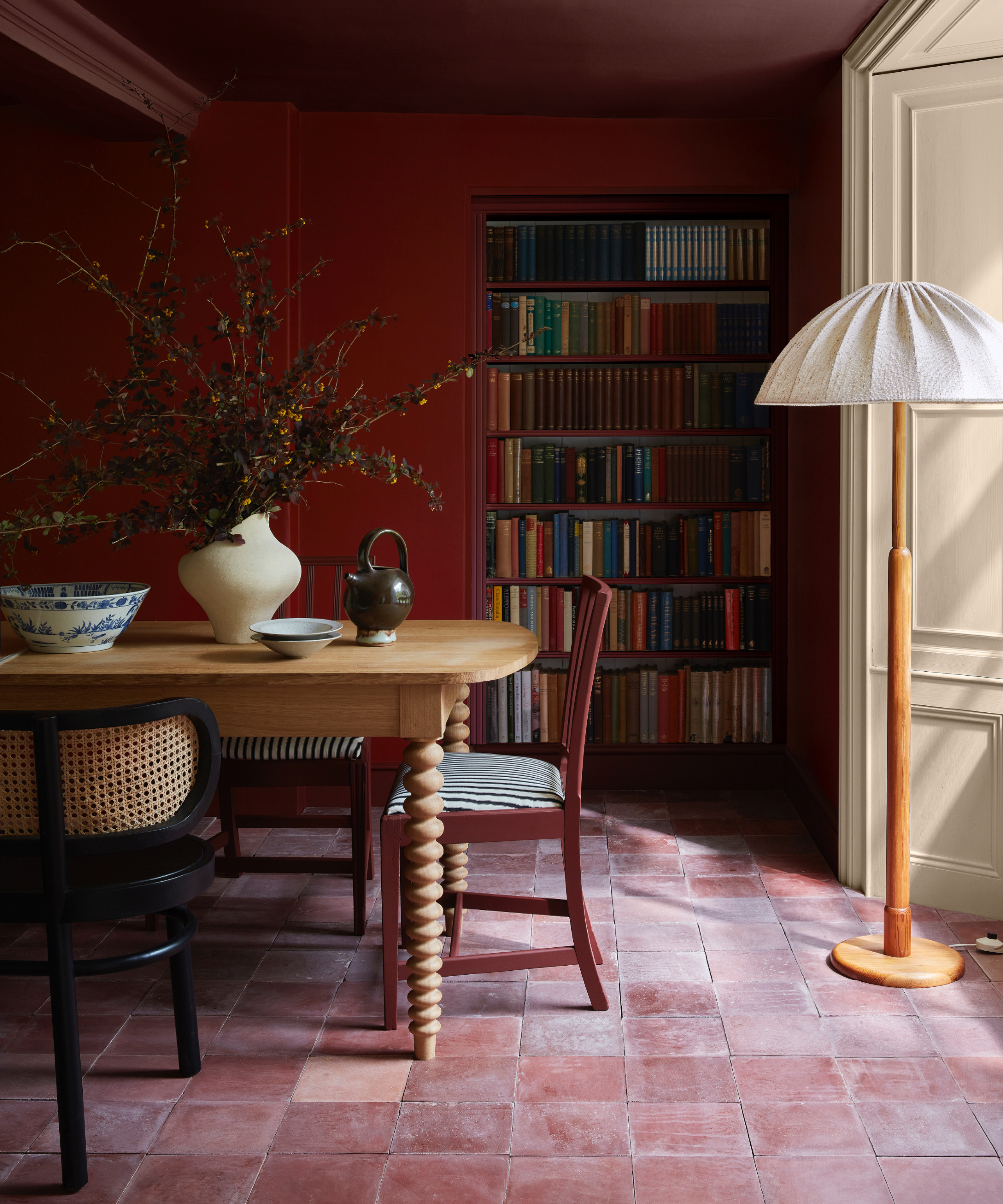 Rich red dining room color soaked, terracotta tiles, floor lamp, wooden table, red and black chairs, bookshelf