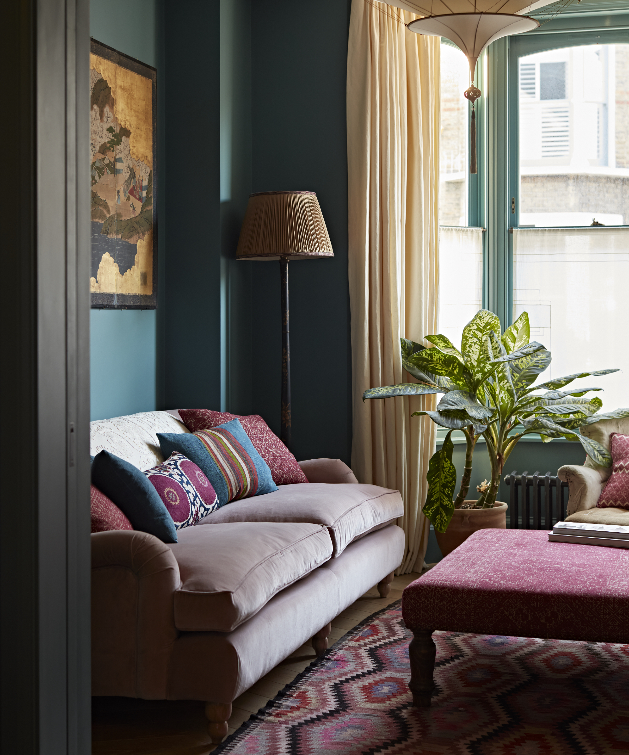 Teal living room with light pink couch, plum foot stop, cream curtains, floor lamp, plant, artwork, ancient style carpet