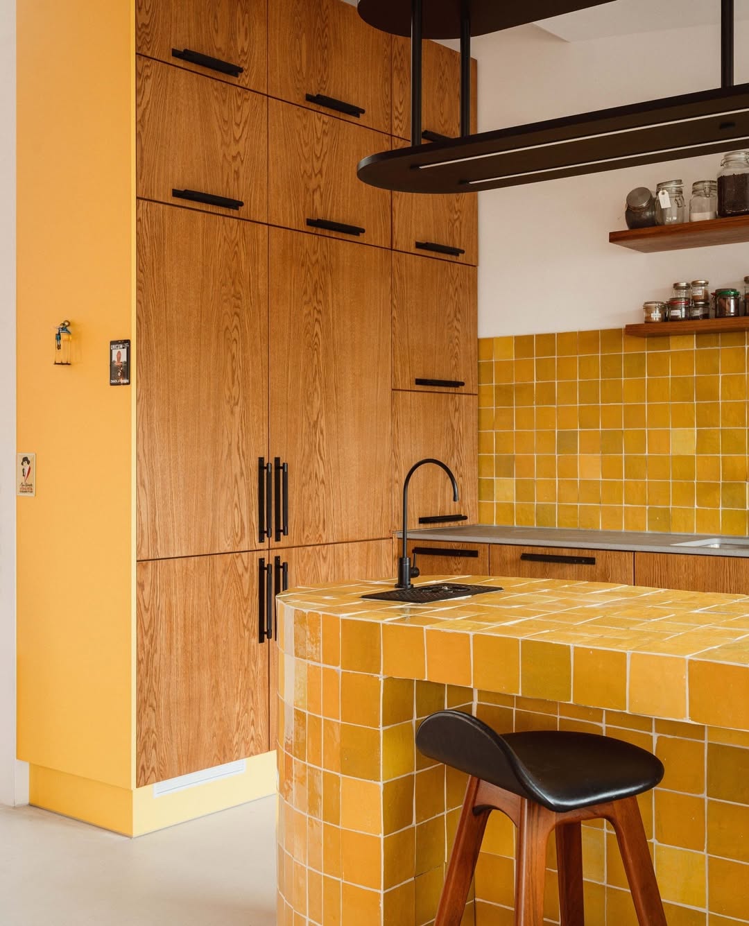 Yellow kitchen with yellow tiled counter, island and back splash, yellow panel in cupboards with wooden finish, black handles, floating shelves