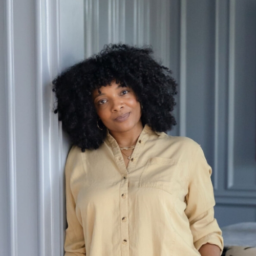 Image of a woman in a yellow button-up shirt. It leans against a light blue wall that forms it. 