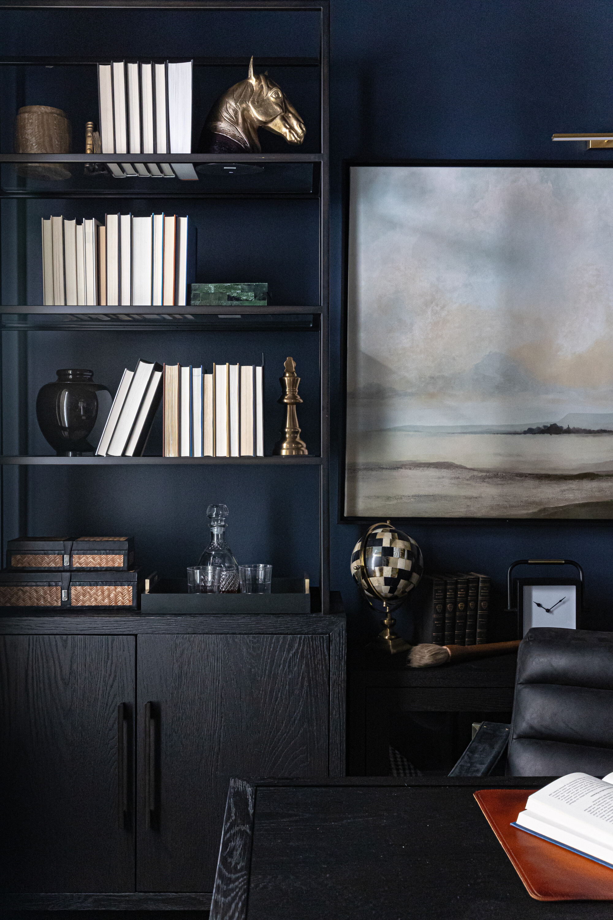 Image of a dark blue room with slim, black shelf and storage device full of books and jewelry. Next to the shelf is a black desk and a chair and a light landscape painting.