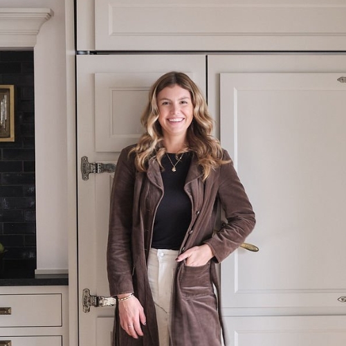 Picture of a woman in a black top, white pants and a long brown coat. It stands in front of cream -colored cupboards.