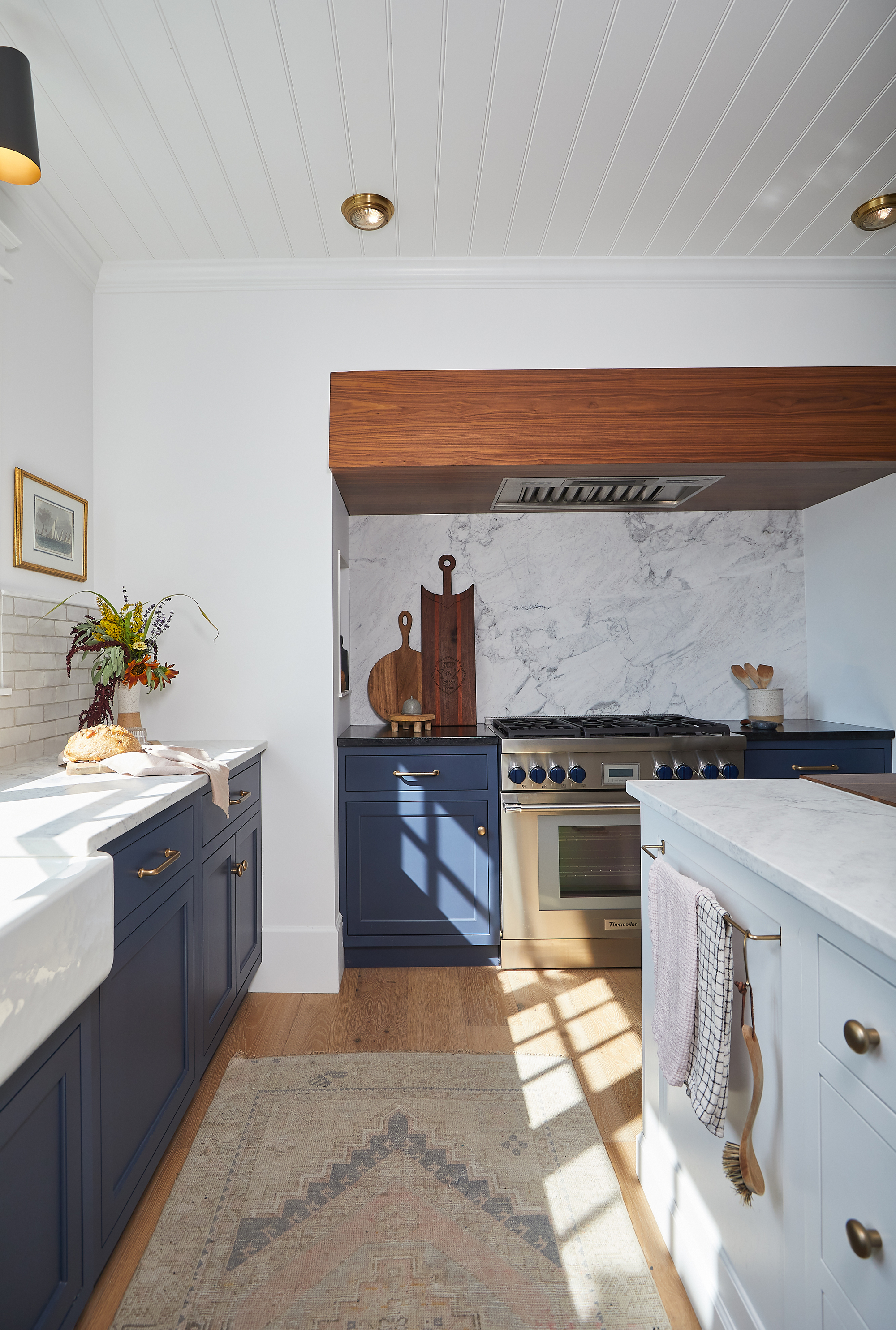 Image of a white kitchen with white marble slabs and back splash. The cupboards are painted in dark blue paint. The floors are hardwood and between the counter and the kitchen island there are a Taner runner carpet.