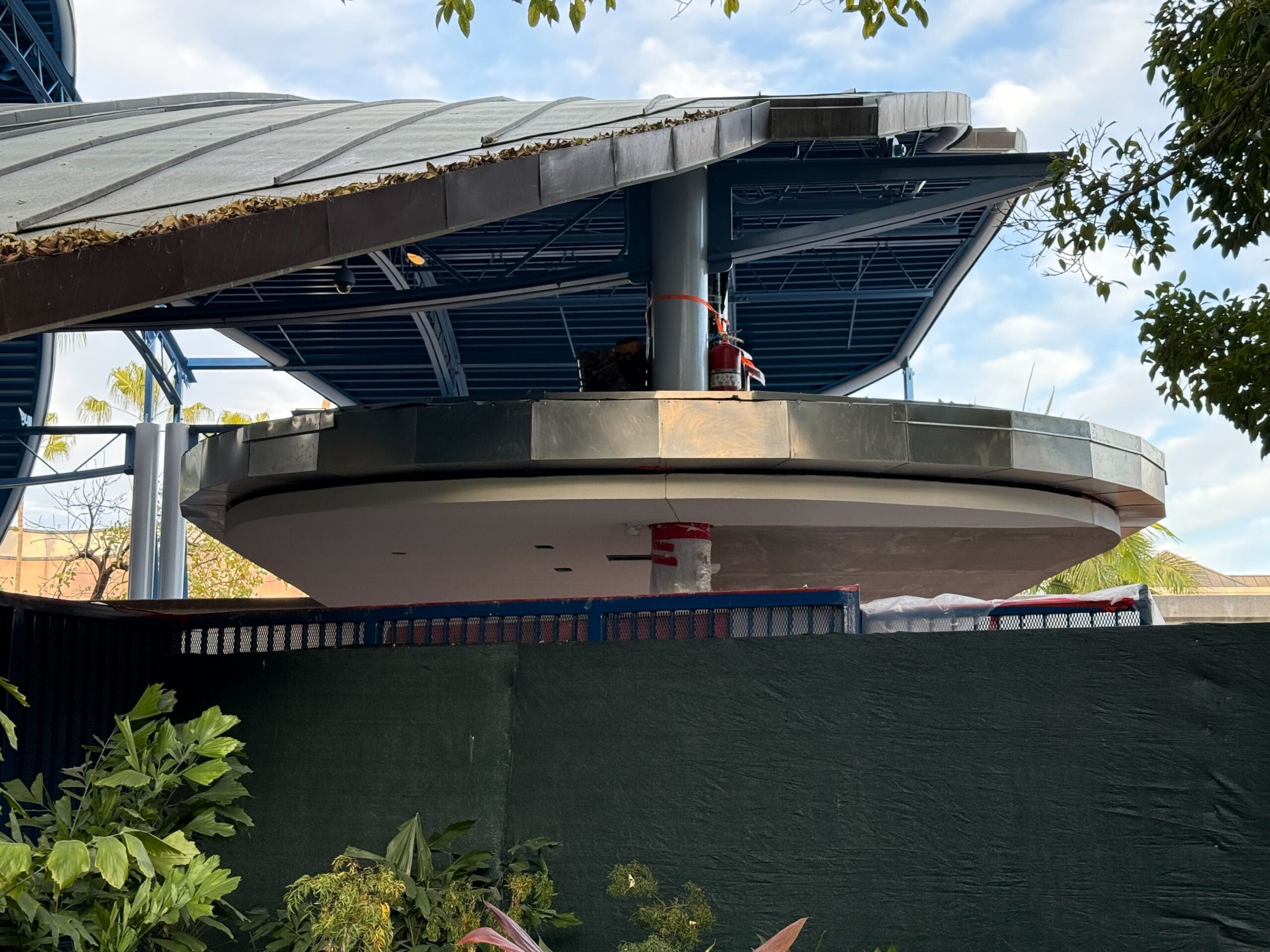 A construction site shows a circular structure with a metal roof that resembles the beginning of a Disneyland monorail station. Green fence and lush leaves offer an enchanting foreground.