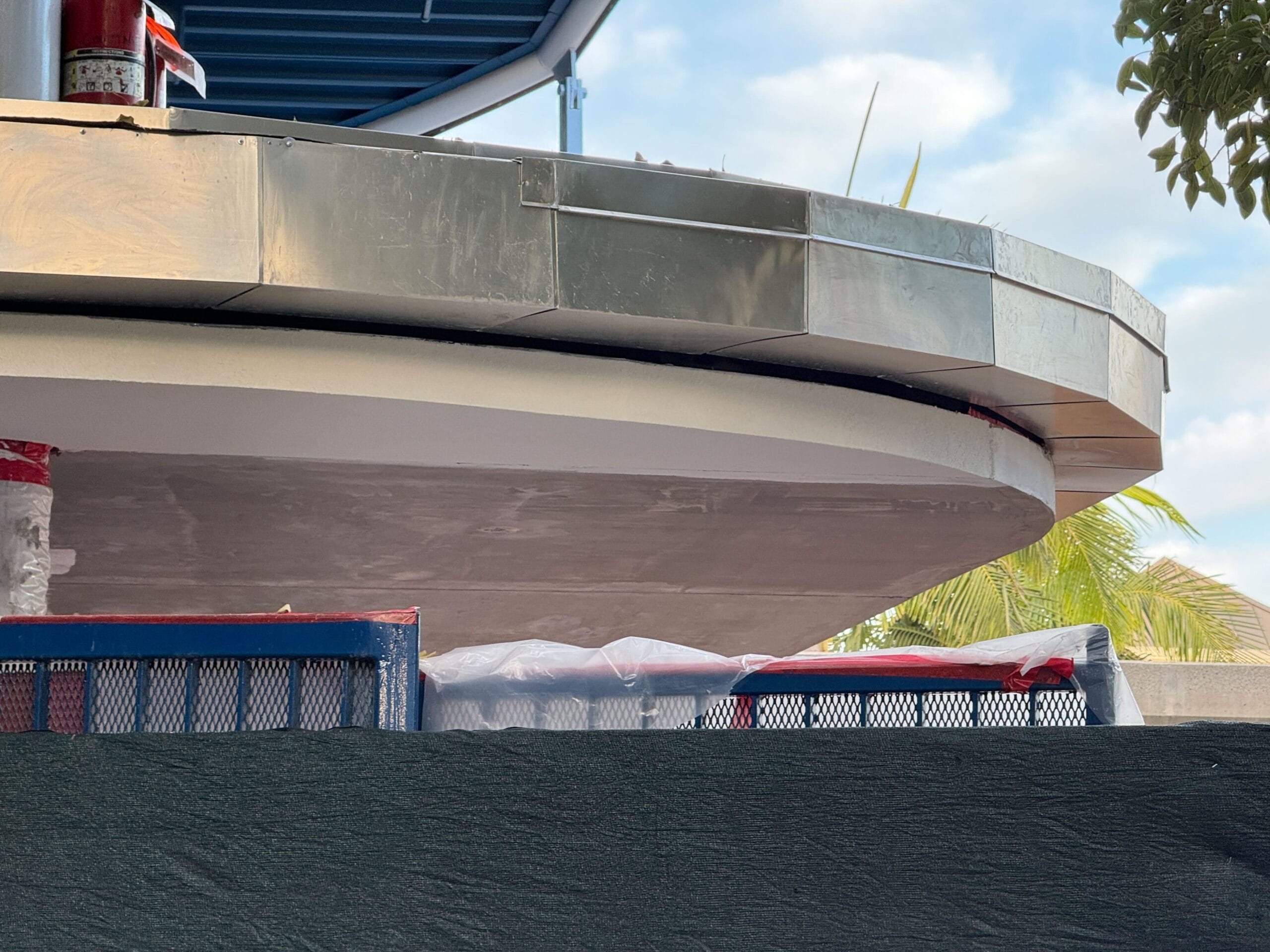 A circular building with metallic equipment is under construction, partly hidden by fences and resembles a future Disneyland monorail station. On the right there is a tree and a blue sky with clouds over it.