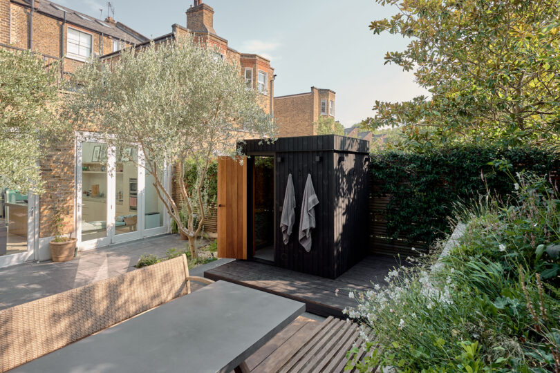 Garden shower outdoors with wood privacy and hanging towels, surrounded by plants and trees, near a brick house with large windows and a terrace table