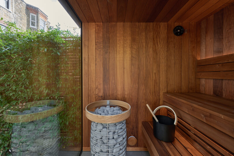 Modern sauna with wooden walls and benches, a metal basket with stones, a wooden trowel and a black bucket. A large window shows green green outside