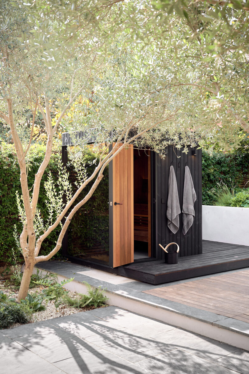 A modern wooden sauna in a garden with open doors and two towels that hang outside. A irrigation box is placed nearby, surrounded by trees and green