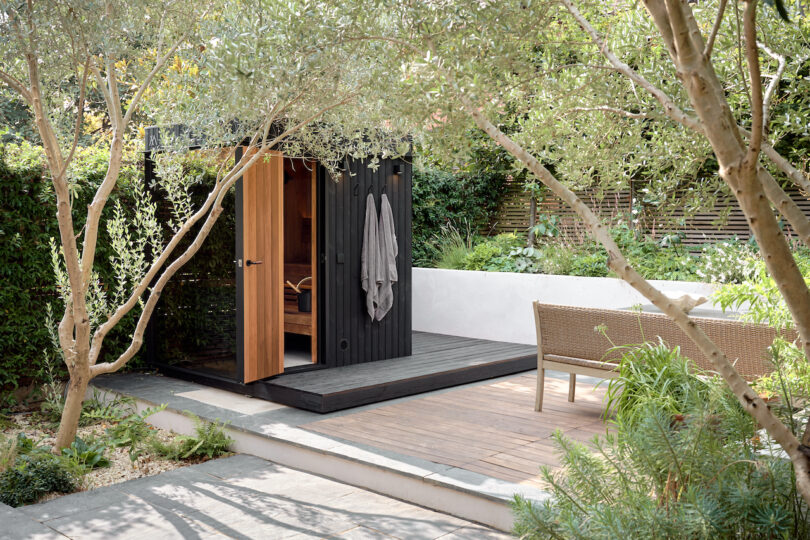 Small wooden sauna with a towel hanging on the door, surrounded by trees and green, next to a basket on a wooden deck