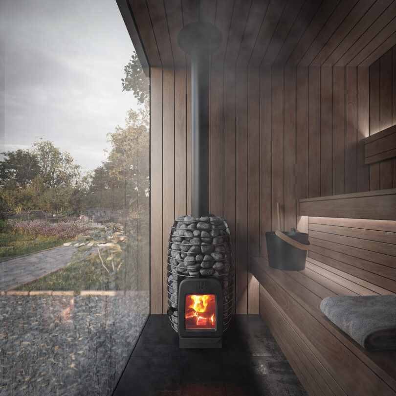 A wooden sauna interior with a stone oven with burning fire. A towel and bucket sit on the bench. A large glass window offers a view of the outdoor landscape