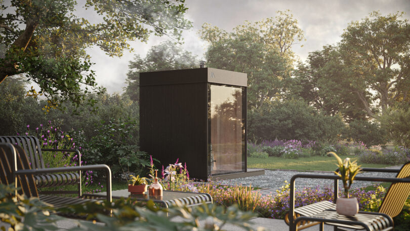 A modern black sauna outdoors is surrounded by lush green and flowering plants. Terrace furniture with potted plants is in the foreground