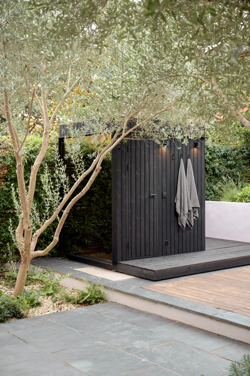 Hölzer sauna outdoors with gray towels, surrounded by green and a tile path in the foreground.