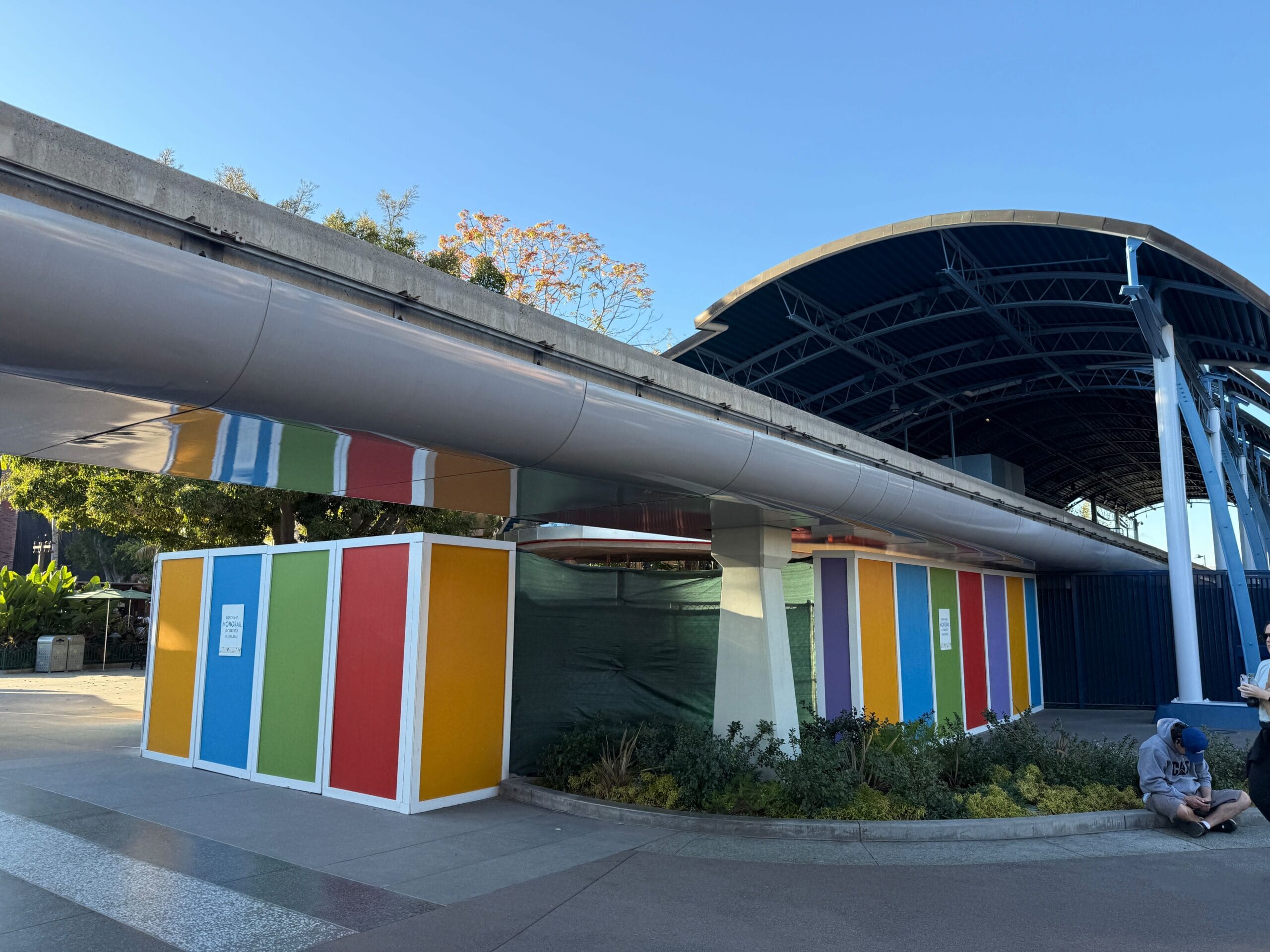 Colorful construction barriers surround a path under a curved roof structure with a clear blue sky.